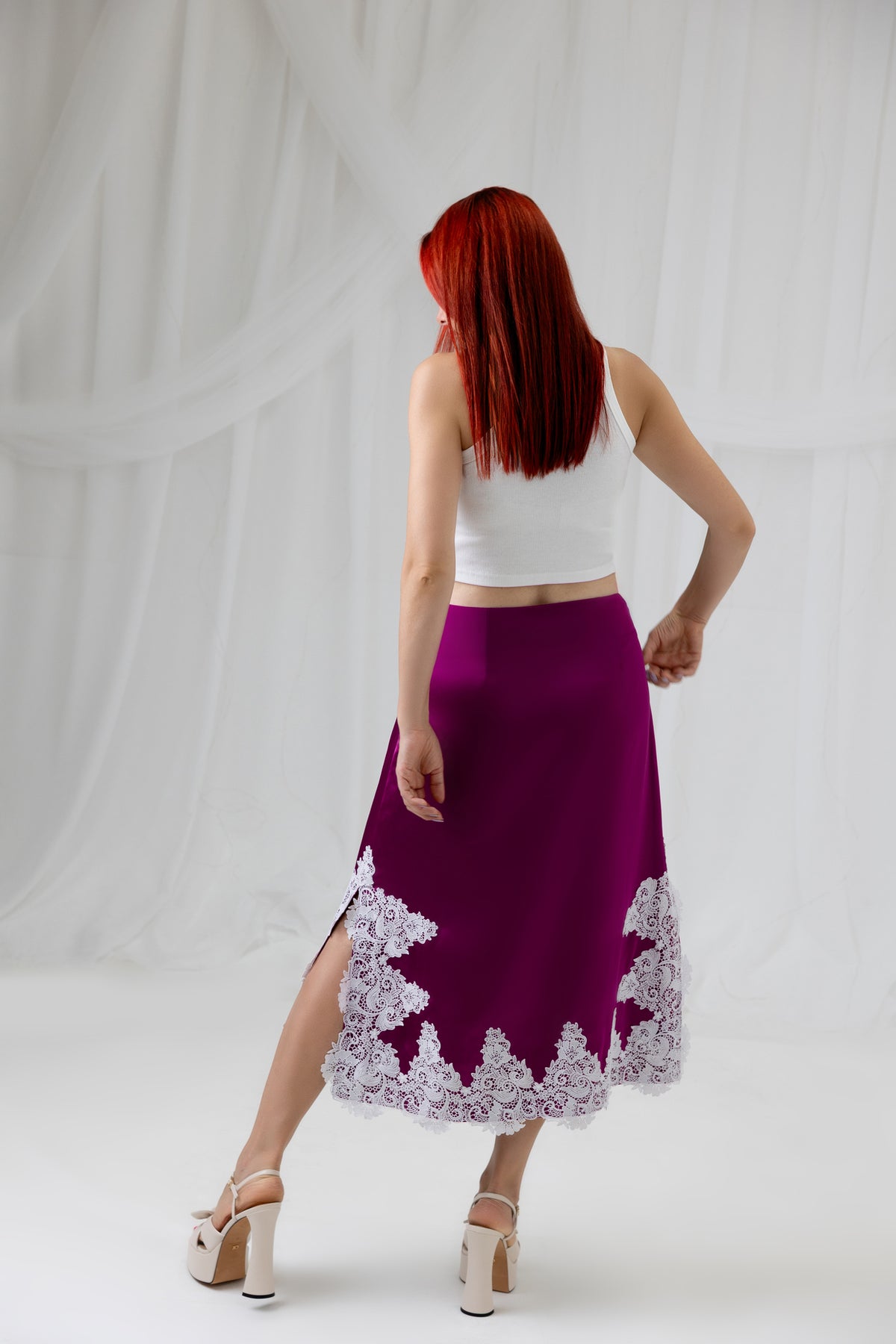 Model wearing the House of Lluvie' Burgundy Lace Whisper satin midi skirt with a white top, back view.